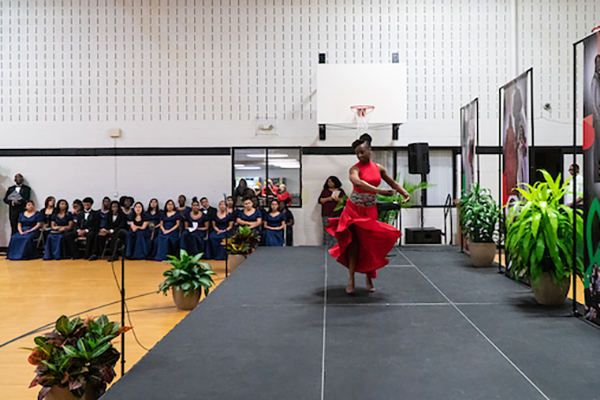 A woman in a red dress dances on a stage set up in a gymnasium. The stage is decorated with vertical banners and potted plants. Against the far wall, women in matching navy blue gowns sit in two rows of chairs.