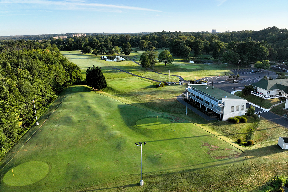 Aerial view of a golf course, with a large patch of bright green grass to the left of a cluster of white buildings and a large parking lot. Several circular, bright green putting greens with flags marking the holes are visible on the course, as well as several small water features. A black asphalt path winds around through the course, which is dotted with and surrounded by trees.