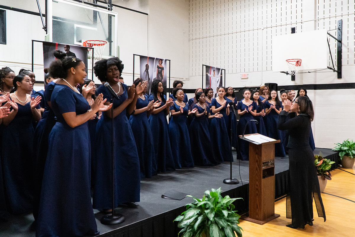 On a stage set up in a gymnasium, a large choir of women in matching navy blue gowns sings and claps their hands. Their choir director stands on the floor in front of them, at a wooden podium, conducting them.