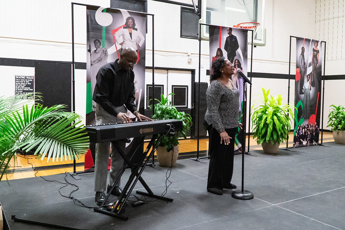 On a stage set up in a gymnasium and decorated with vertical banners and potted plants, a woman sings at a microphone while a man stands next to her playing a keyboard.
