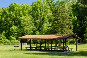 A picnic shelter in a grassy lawn with four picnic tables under it, on a concrete patio. There is a grill installed in the grass nearby. Past the far edge of the grass are tall trees.