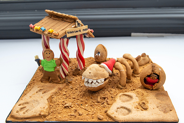 A gingerbread man under a candy cane stand. He is standing on the gingerbread dinosaur park, and has found a giant festive gingerbread dinosaur fossil wearing a Santa hat.