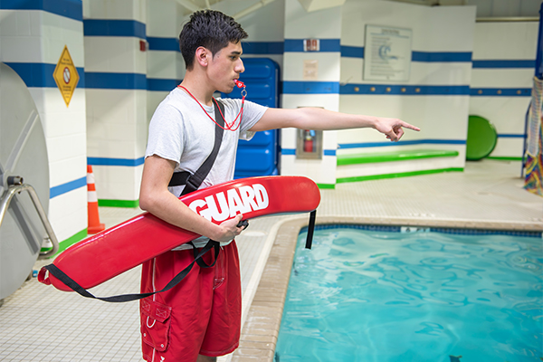 At an indoor pool, a lifeguard standing on the deck wearing red shorts and a white shirt and holding a red flotation device blows his whistle while pointing into the pool.