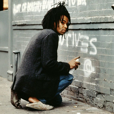 A man looks up furtively while crouching on a sidewalk next to a brick building covered in white graffiti. In his hand, he holds a can of spraypaint pointed at the wall.