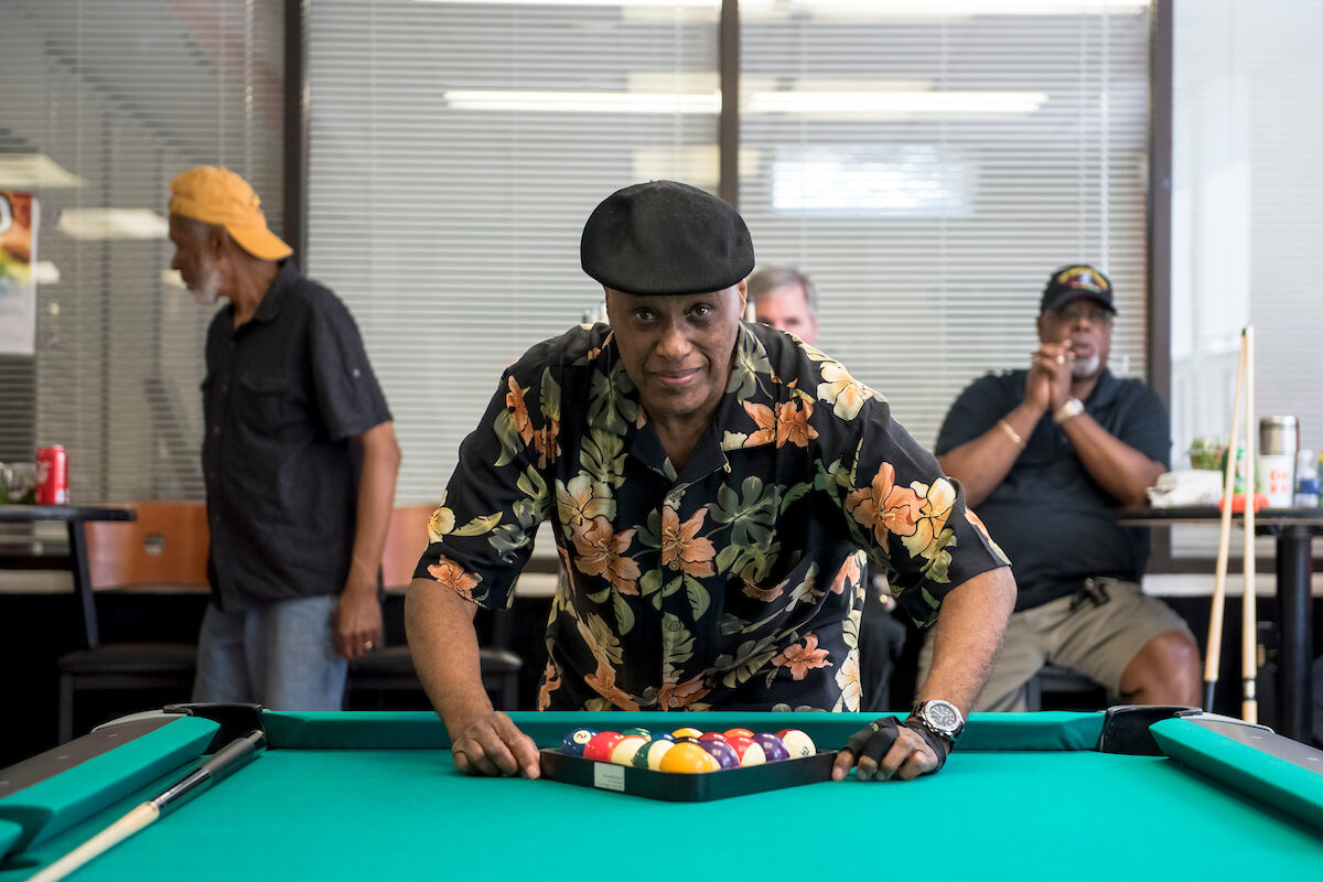 A man in a black cap and a floral shirt racks the balls on a billiard table. Several men wait behind him with pool cues.