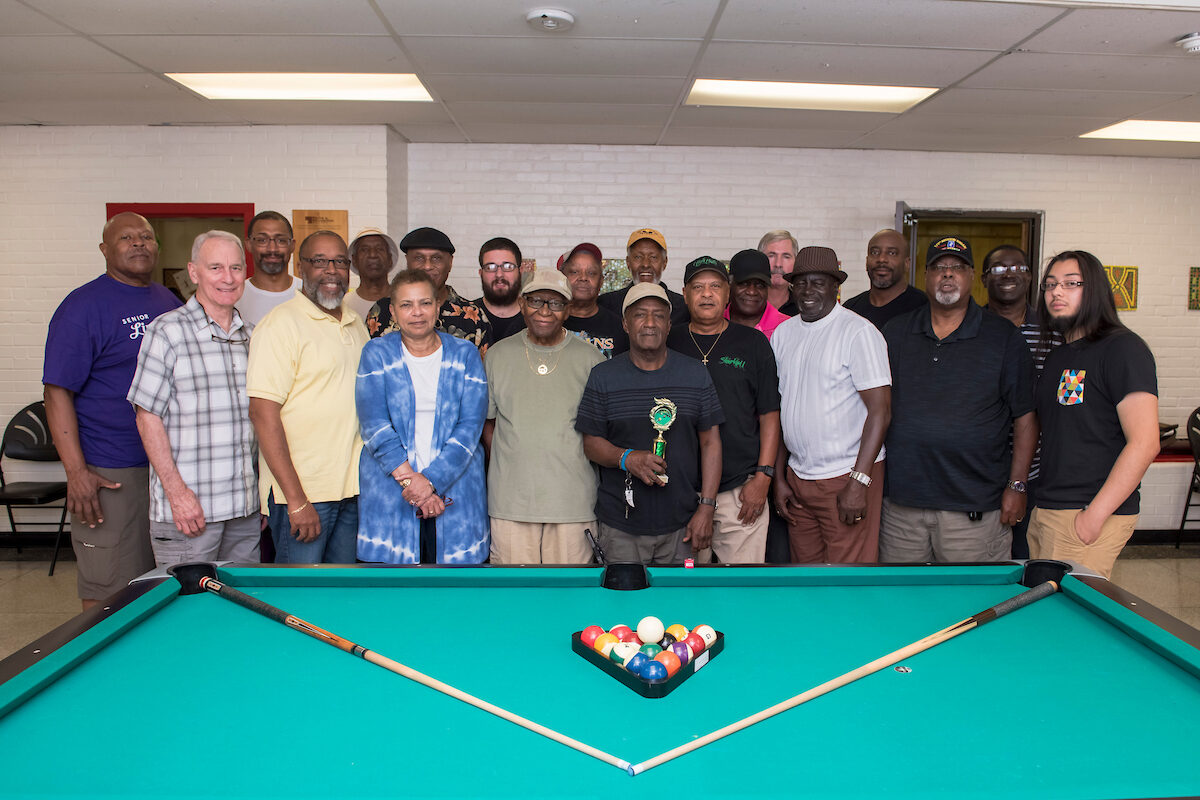 About 20 men stand close together, posing for a photo behind a billiard table, which is artfully arranged with a rack of balls and two angled pool cues.