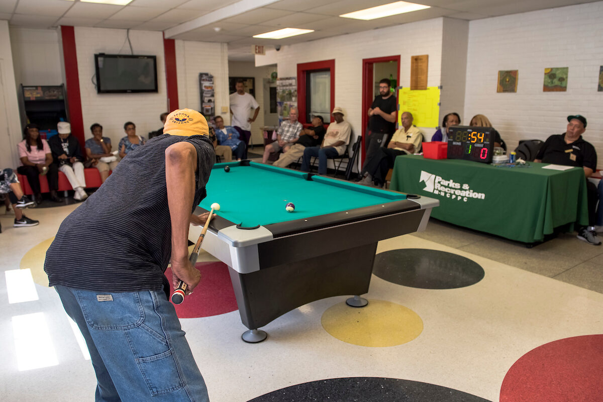 A man bends forward to take a shot at a billiard table. Spectators line the far walls. To the right rear is a long table with a green Parks and Recreation tablecloth and a small, lighted score-board. The linoleum floor has a polka-dot pattern.