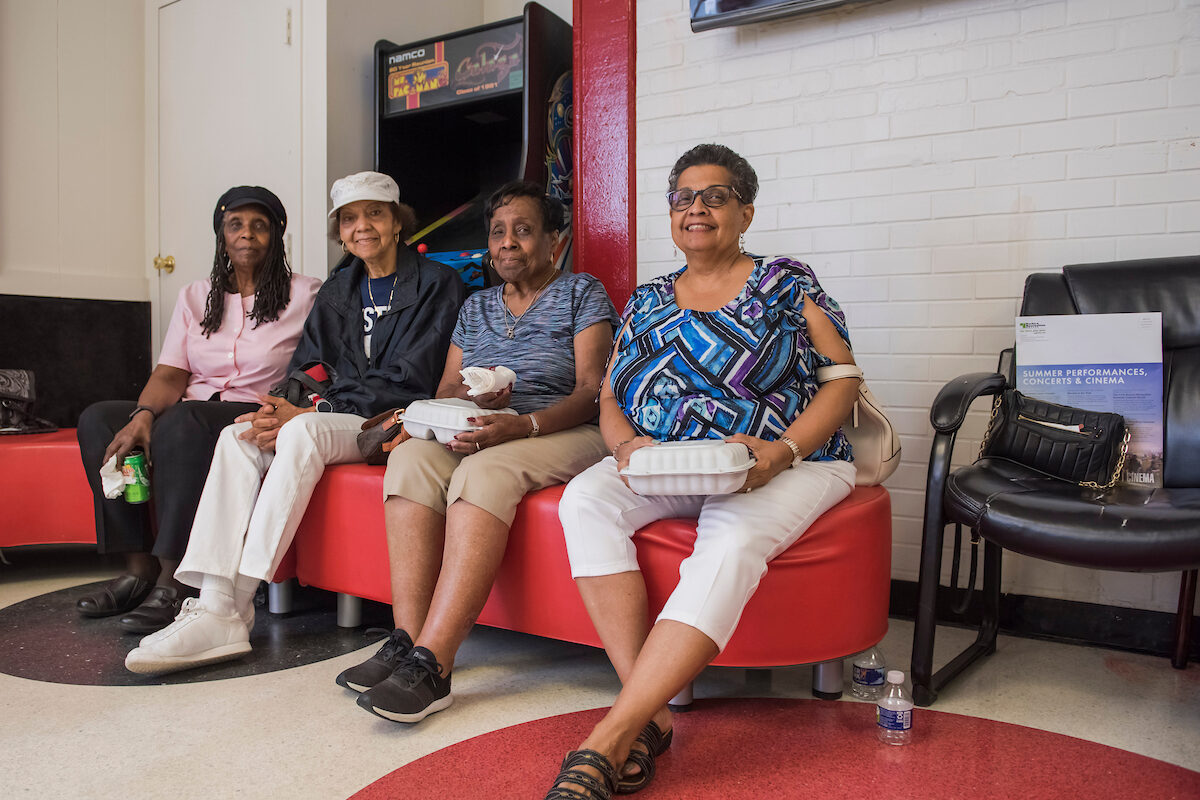 Four women sit on red, upholstered, kidney-bean-shaped bench in a game room. Behind them is a classic arcade game. The linoleum floor has a decorative polka-dot pattern.