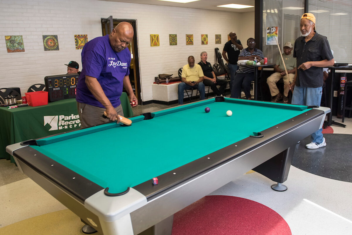 A man in a purple shirt brushes the felt on a green billiard table. Behind him is a long table with a green Parks and Recreation tablecloth with a small, lighted scoreboard on it. Spectators line the far wall.