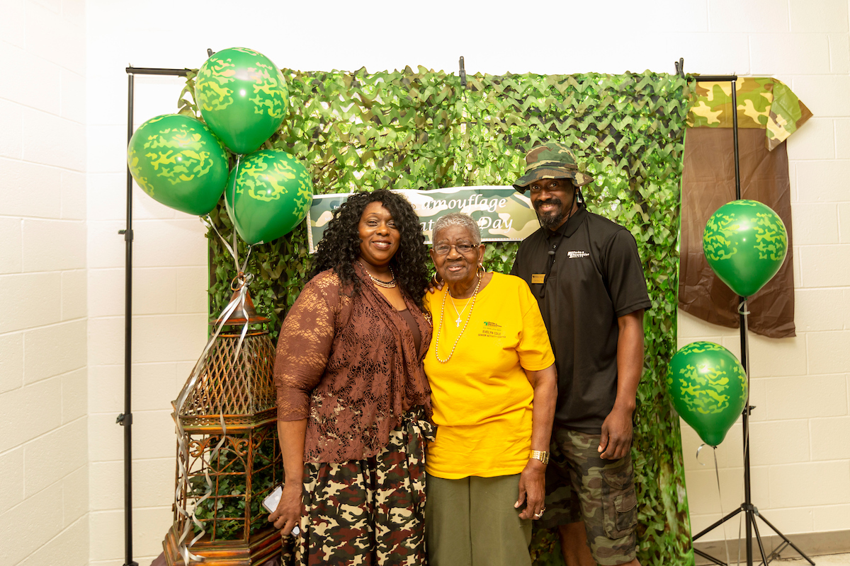 Three adults pose between green balloons in front of a leafy, camouflage-decorated backdrop. The woman on the left wears a camouflage skirt; the man on the right wears a camouflage hat and shorts.