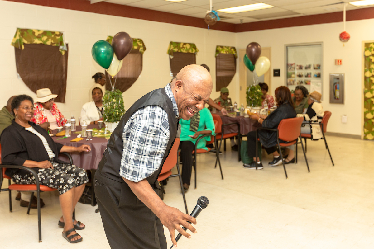 A singer wearing a vest and holding a microphone stands in a party room near several tables of seated adults. The room is decorated with balloons and camouflage banners.