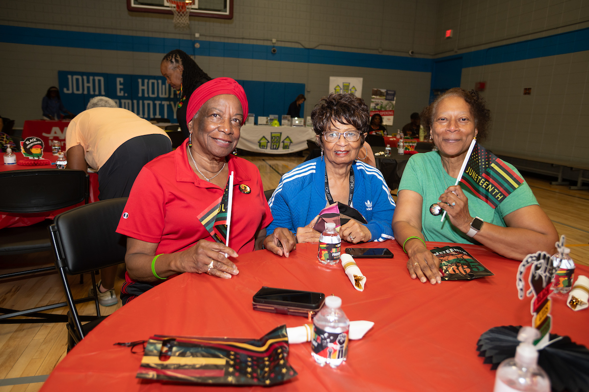 In a gymnasium set up for a party, three smiling women pose while sitting at a party table with a red tablecloth. They hold small Juneteenth flags.