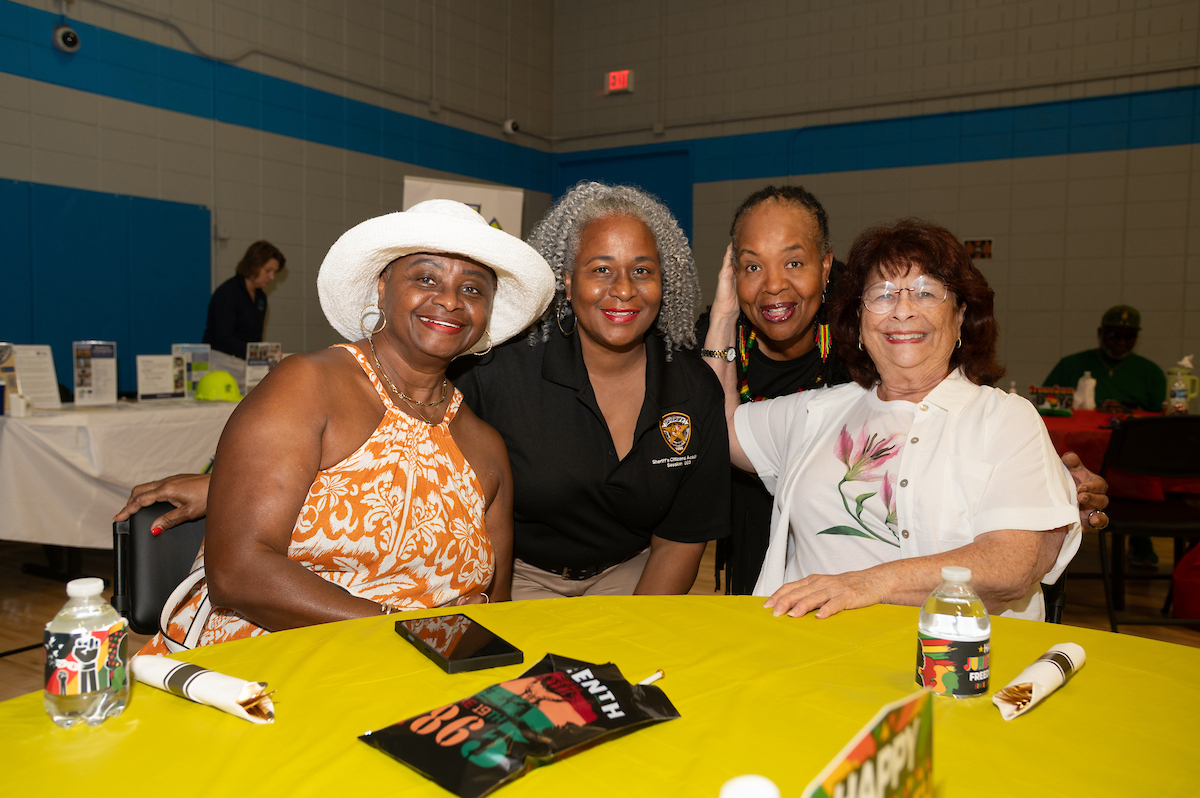 Four smiling women pose for a picture at a party table with a yellow tablecloth. Promotional items on the table are decorated with Juneteenth colors and messaging.