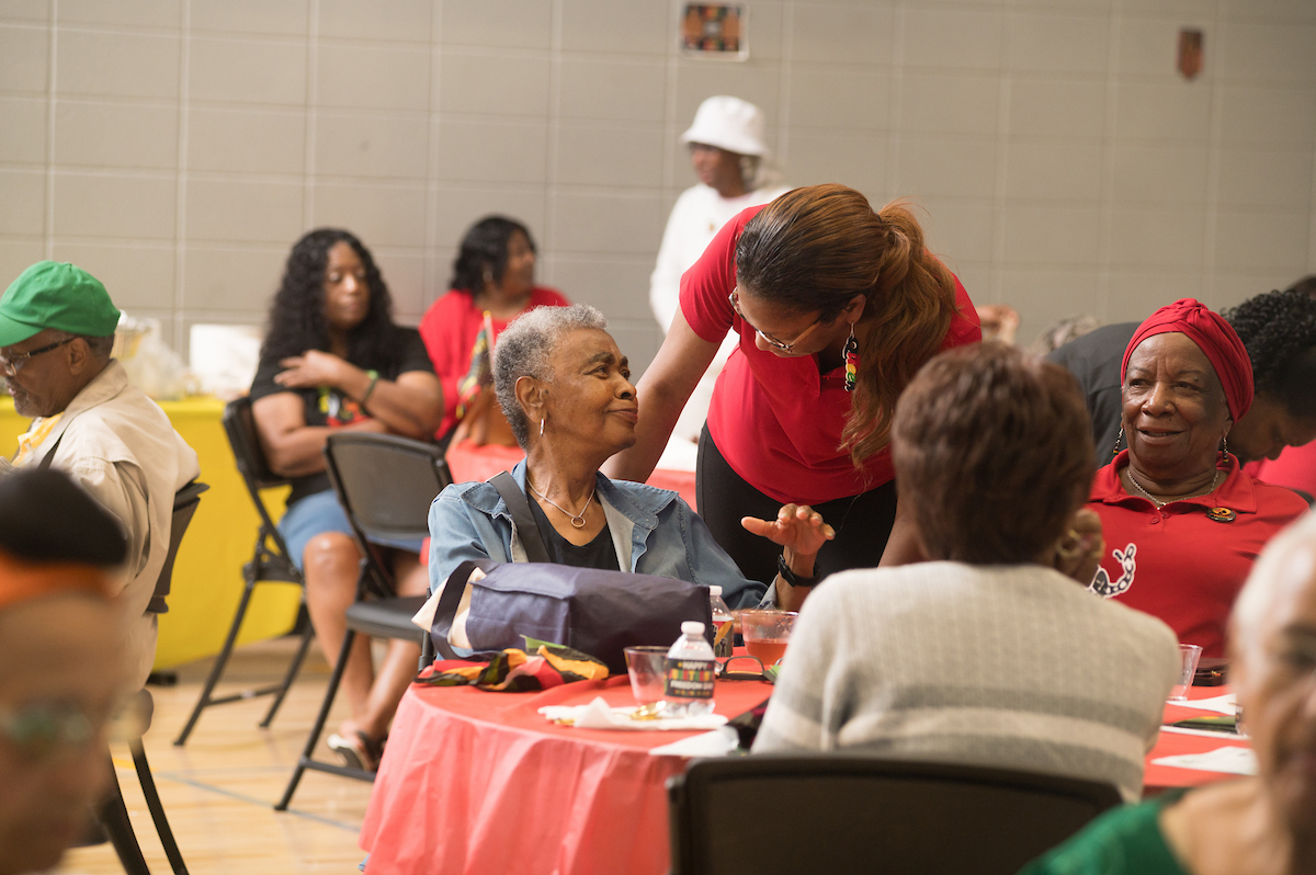 A staff member in a red shirt bends down to speak to a smiling woman who is seated at a table in a gymnasium with other guests. The tables have festive red tablecloths.