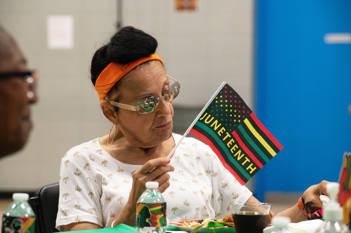 A woman with an orange headband sits at a table holding a miniature flag with red, black, yellow, and green stripes that says JUNETEENTH. Water bottles on the table are also decorated with a Juneteenth theme.