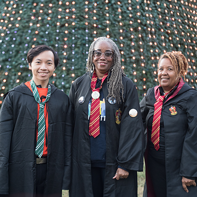 Three people dressed as wizards standing in front of a tree adorned with strings of white lights