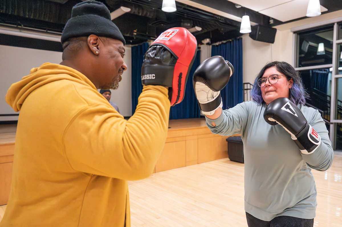 Inside a dance-type studio, a young woman with purple streaks in her black hair wears boxing gloves and aims a right-handed punch toward her coach. He holds one hand up near his face with a red pad on it for his student to punch.