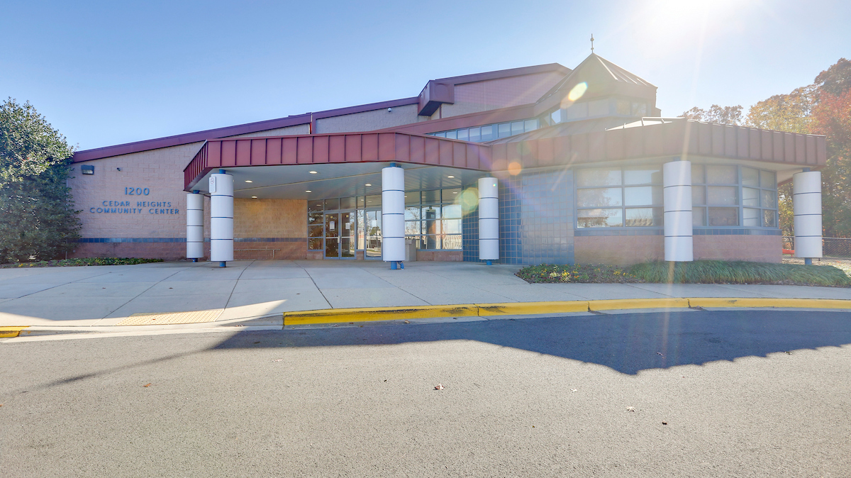 Sunny exterior view of a large brick, metal, and glass building with a red roof and a red portico over the entrance supported by white columns. Blue letters on the building read "12OO CEDAR HEIGHTS COMMUNITY CENTER."