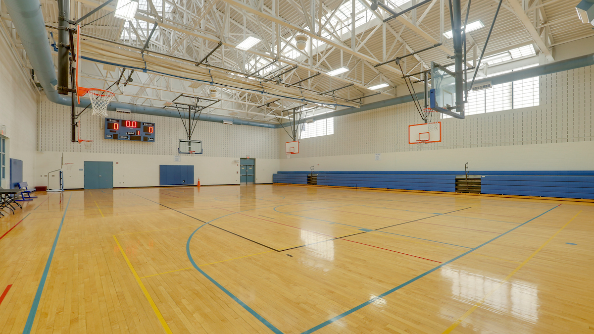 A gymnasium with a shiny wooden floor and multiple basketball goals suspended from the ceiling. The room is white with blue trim and blue bleachers along the far wall.