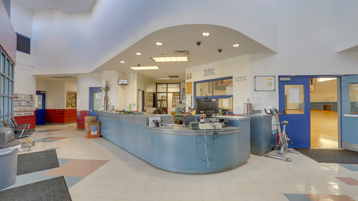 A long, curved, blue front desk in a high-ceilinged lobby area just inside the entrance. To the right of the desk are blue double doors to a gymnasium; to the left of the desk is a hallway with red tiled walls.