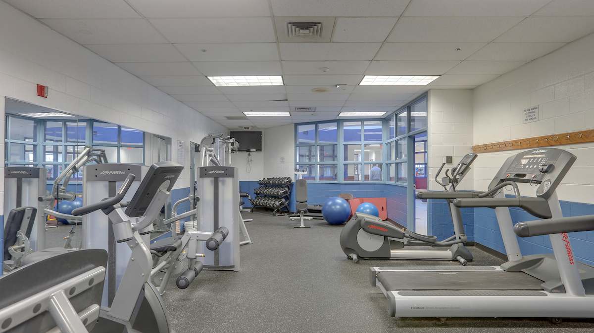 A fitness room with blue and white walls and a bank of windows through which can be seen a hall/lobby area. The room has one mirrored wall and contains treadmills, free weights, exercise balls, a TV, and other various pieces of exercise equipment.