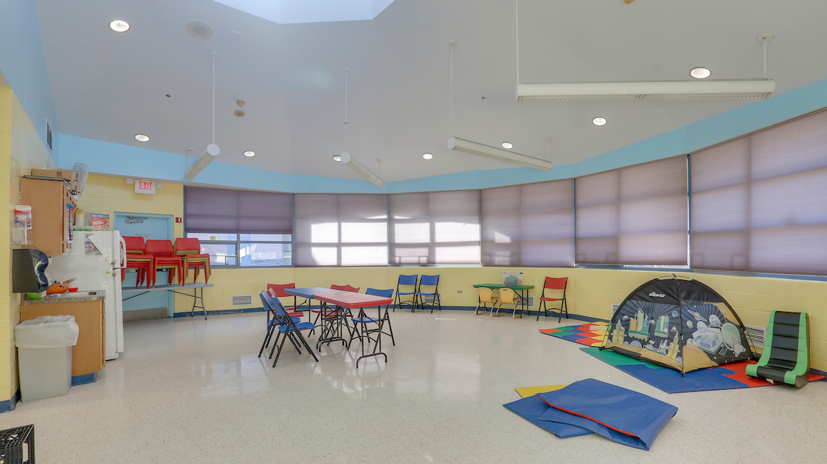 A large circular room with windows along the curved exterior wall covered by purple blinds. The room contains colorful plastic tables and chairs, multiple play mats, a gaming chair, and a colorful tent.