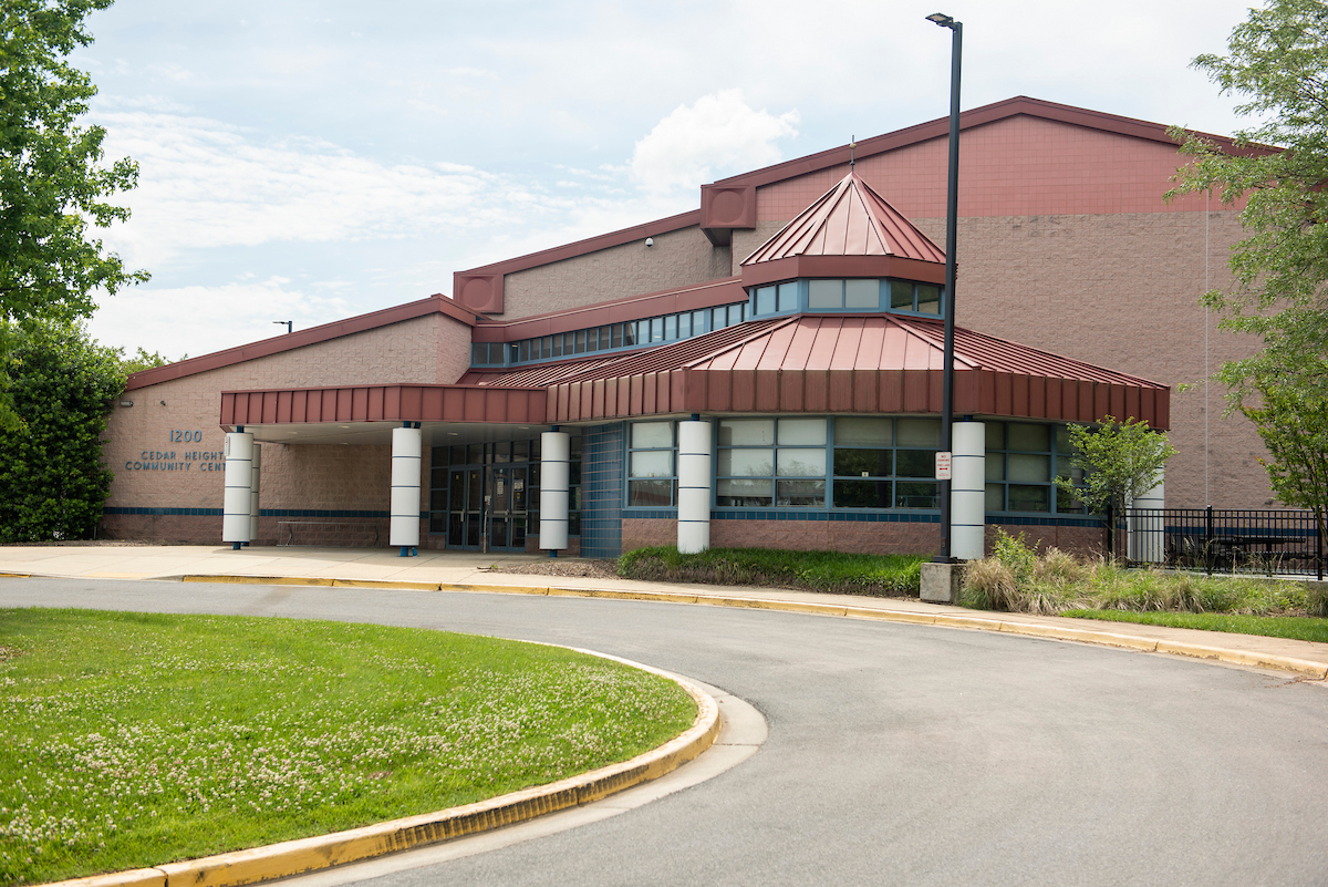 Exterior view of a large brick, metal, and glass building with a red roof and a red portico over the entrance supported by white columns. Blue letters on the building read "12OO CEDAR HEIGHTS COMMUNITY CENTER."
