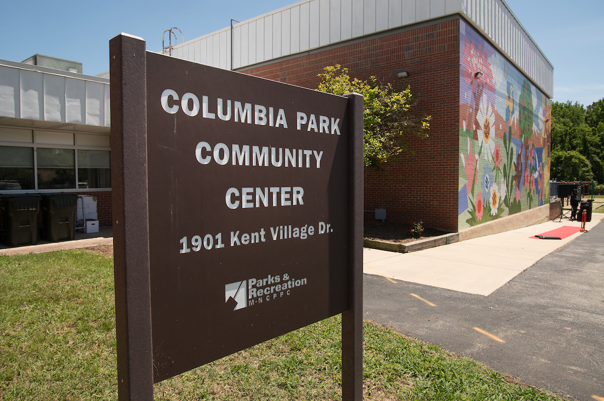 A brown sign reading COLUMBIA PARK COMMUNITY CENTER is mounted in the grass in front of a large, brick and glass building. One wall of the building is covered by a colorful mural.