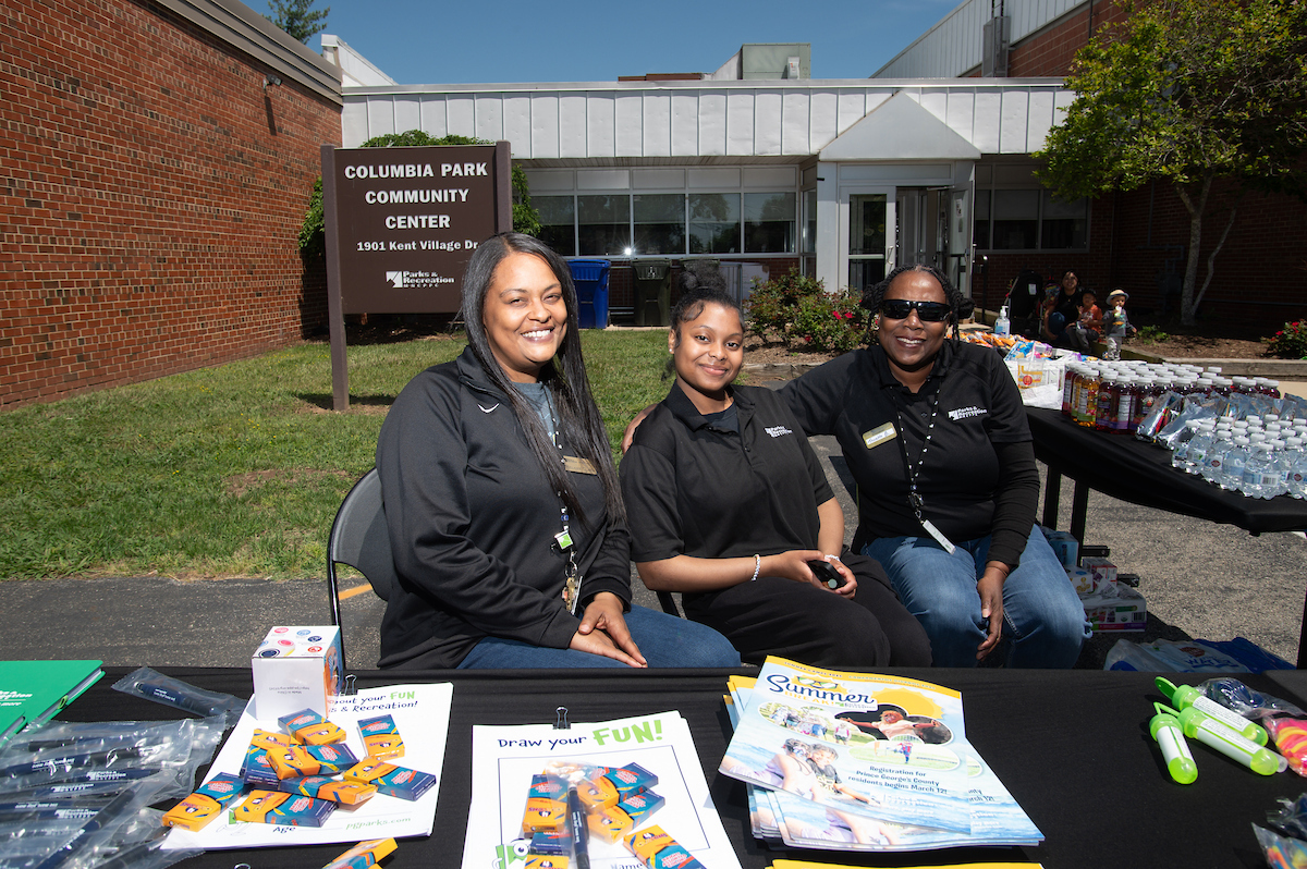 On the grass in front of a brick and glass building, three women in black staff shirts smile while sitting behind a table containing crayons and coloring pages. Behind the women is a brown sign that reads COLUMBIA PARK COMMUNITY CENTER. Another table full of water bottles, juice, and snacks is to the right.