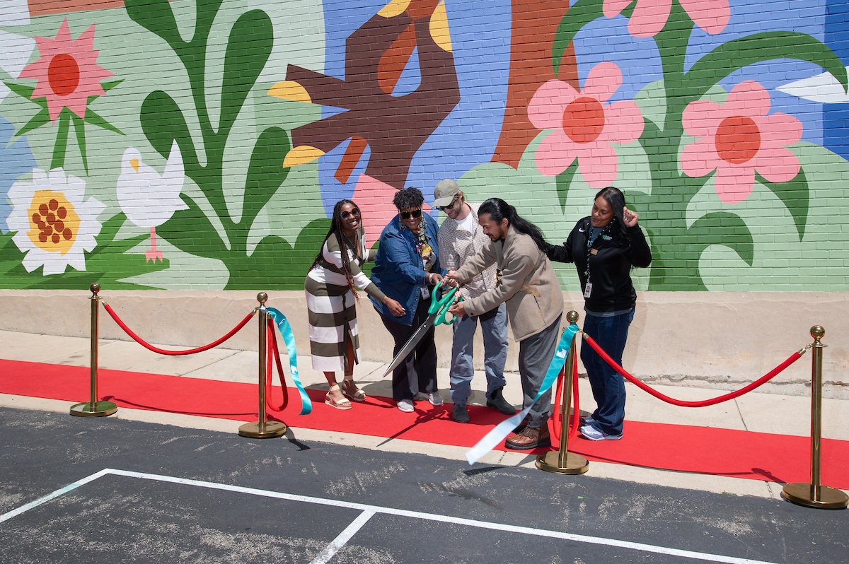 Five adults stand on a red carpet in front of a large mural, watching a man with long black hair use giant novelty scissors to cut a turquoise ribbon attached to brass stanchions. They smile and make celebratory gestures.