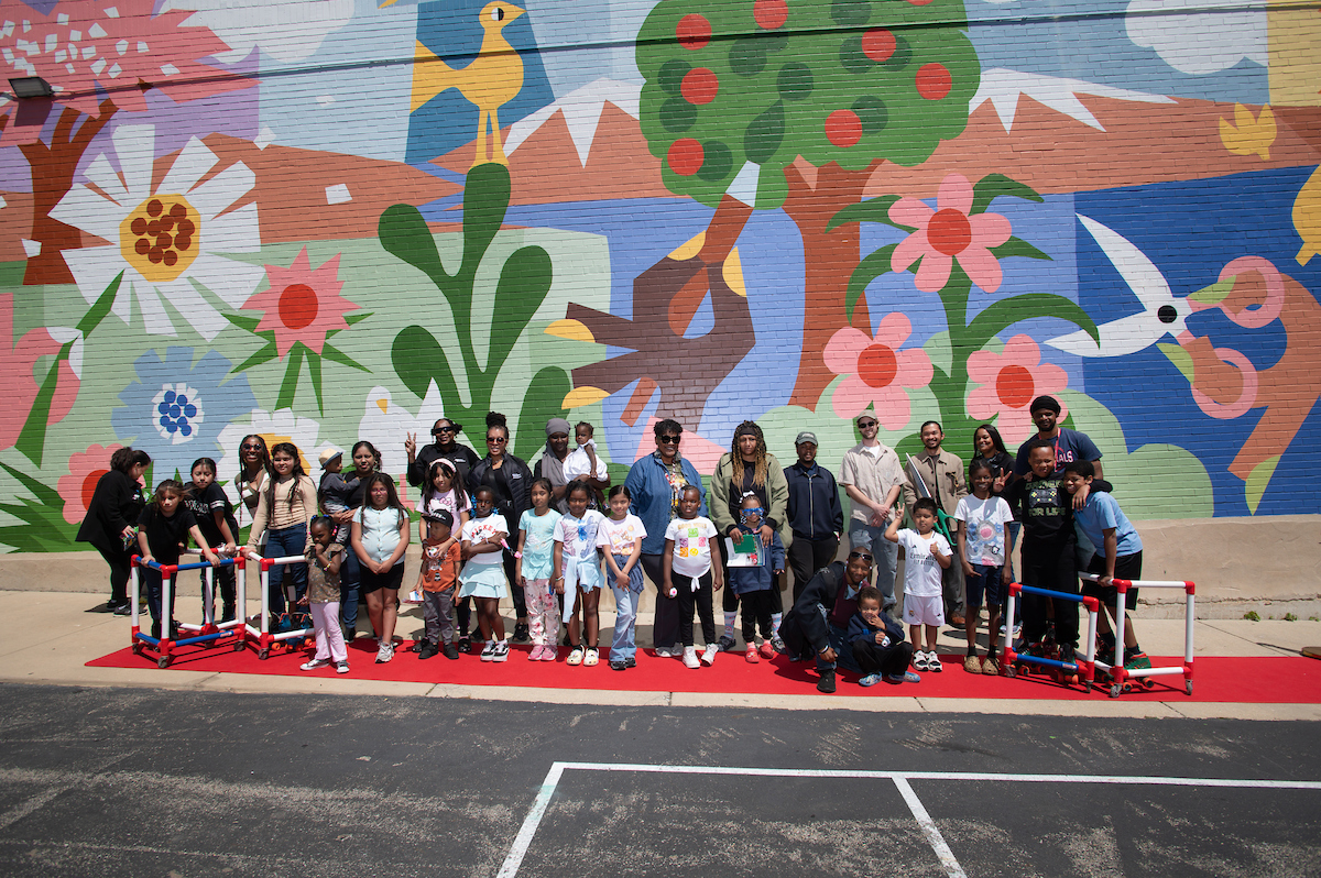 About 40 adults and kids pose on a red carpet in front of a giant colorful mural painted on a brick building. Some of the children wear roller skates and hold skating support frames.
