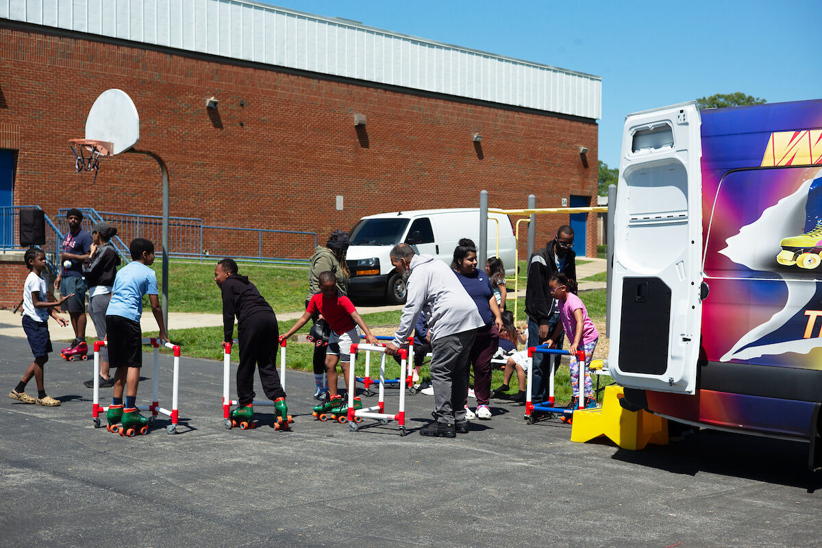 Children roller skate on an asphalt basketball court next to a large brick building. Some hold onto frames on wheels for support.