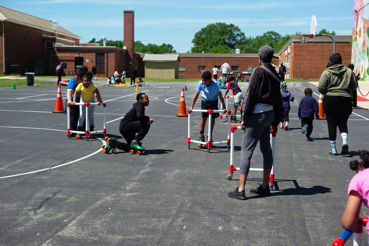 Children roller skate on an asphalt basketball court next to a large brick building. Some hold onto frames on wheels for support.