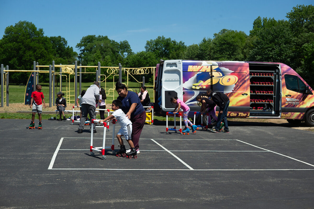 Children roller skate on an asphalt blacktop adjacent to a playground. A van full of roller skates and equipment is parked nearby.
