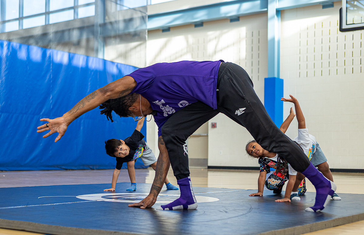 In a gymnasium, an instructor in a purple shirt demonstrates a wrestling position to three small children, who copy his position (both feet and one hand on the wrestling mat, one arm outstretched). The students watch the instructor closely.