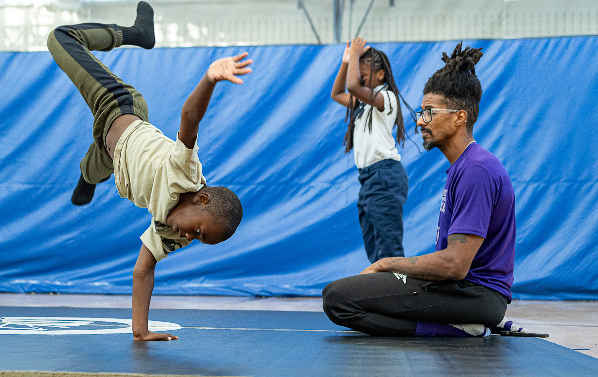 In a gymnasium, a boy in sweatpants does a one-armed handstand-type move on a wrestling mat while his instructor kneels nearby, watching.