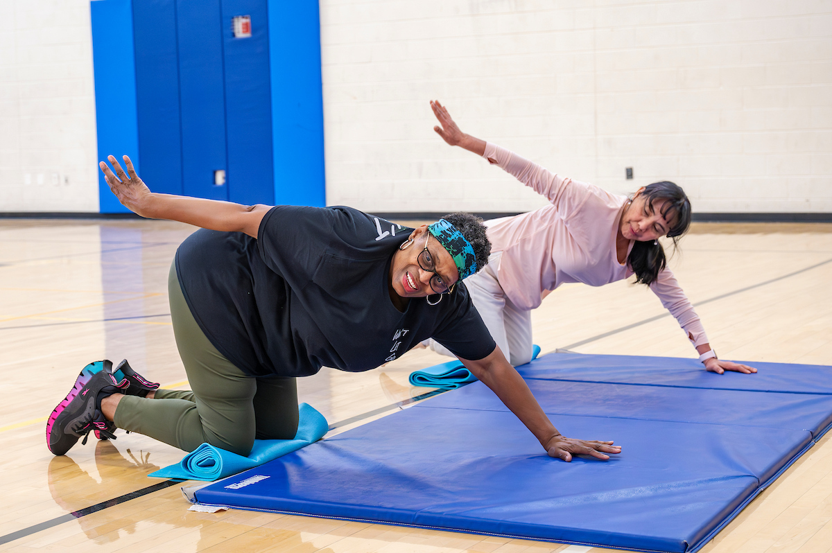 Two women in a gymnasium kneel on rolled yoga mats while supporting themselves with one arm on a blue gymnastics-style mat on the floor. They are executing a stretch.