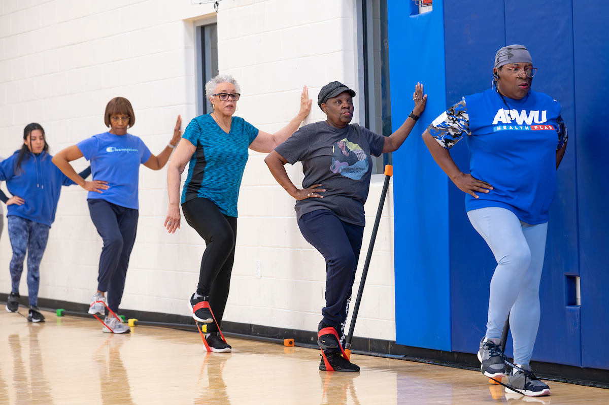 Five women wearing various shades of blue stand along the wall of a gymnasium, bracing themselves with one arm while stretching resistance bands with their feet.