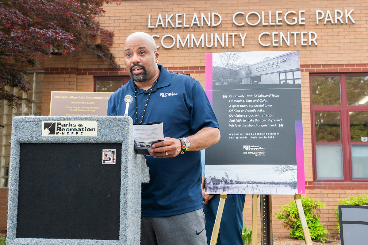 A man speaks at a gray podium outside in front of a brick building with letters that read LAKELAND COLLEGE PARK COMMUNITY CENTER. Behind the man is a poster on an easel featuring a poem and an older, black-and-white photo of the same building.