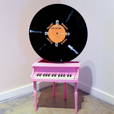 A small pink piano sitting in a corner with white walls and concrete flooring. There is a record positioned on top of the piano.