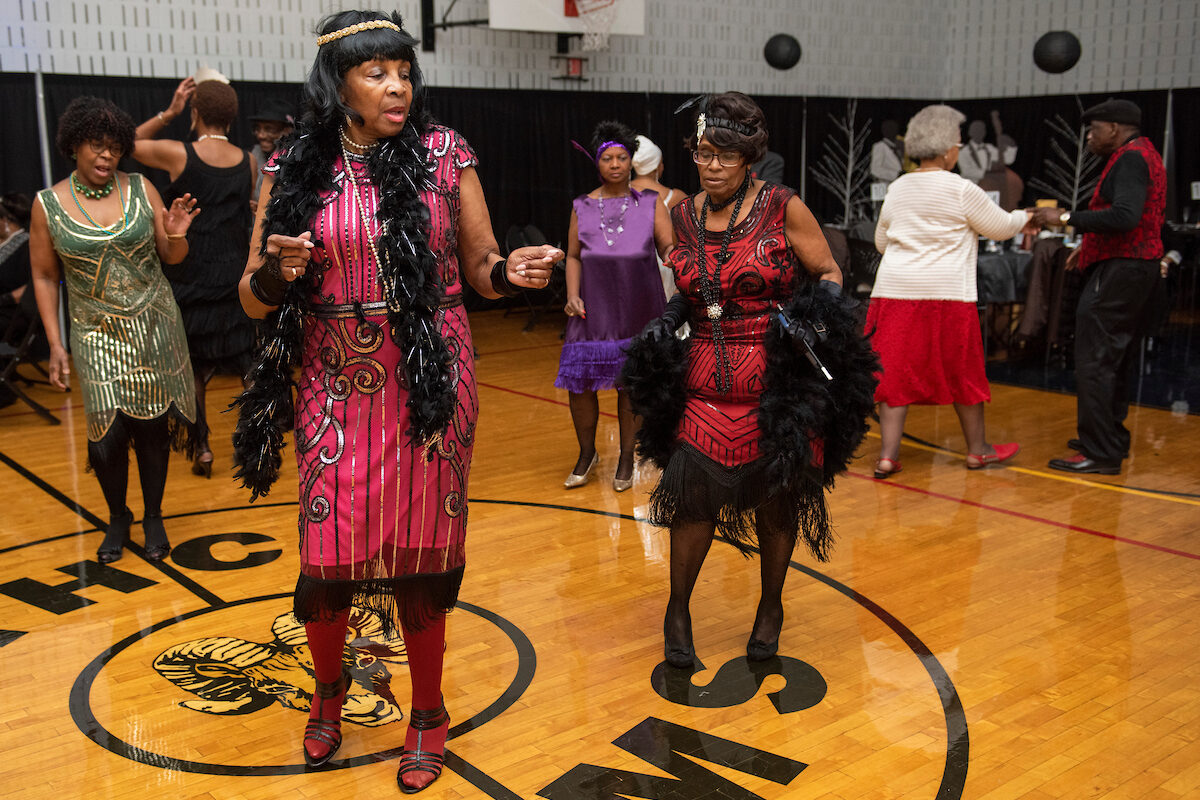 In a decorated gymnasium, seniors in elaborate, beaded 1920s flapper dresses dance. Many wear feather boas, 1920s headbands, and long beaded necklaces.