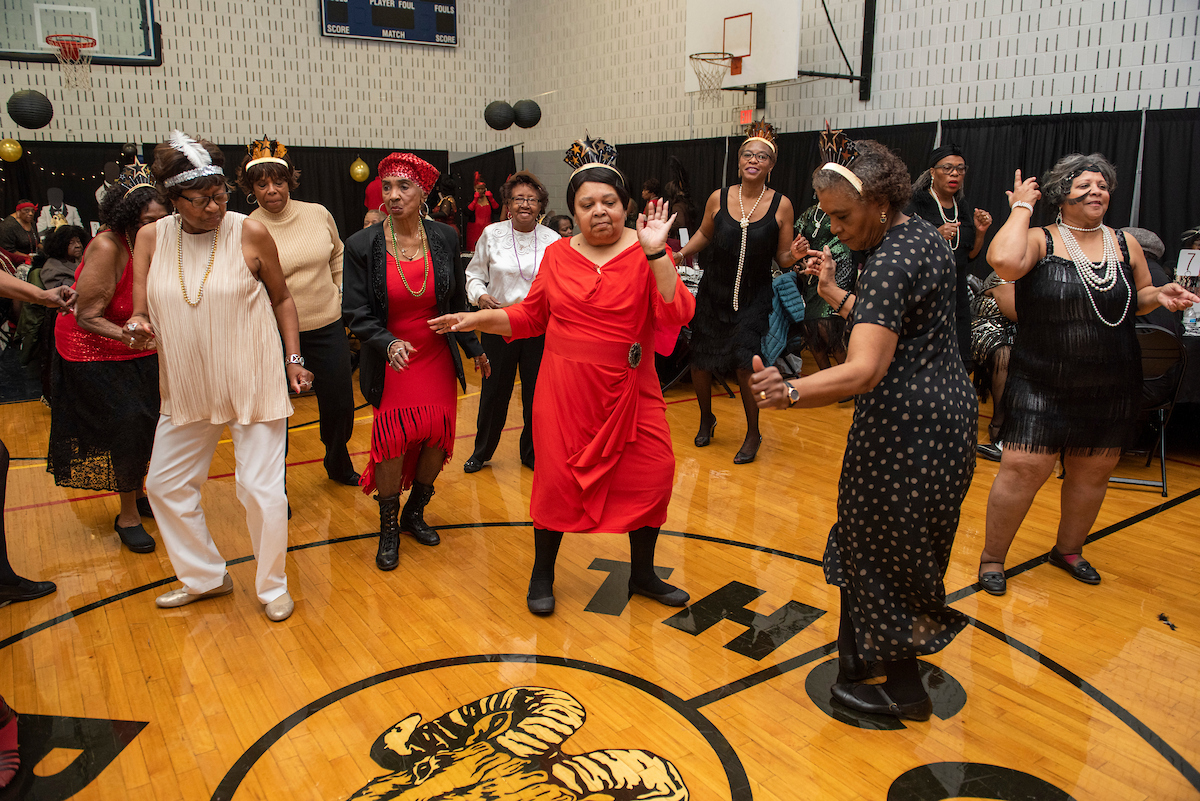 In a gymnasium decorated with balloons and garlands, seniors in 1920s party dresses dance and celebrate. Many are wearing glittery paper party hats of the style worn at New Years Eve parties.
