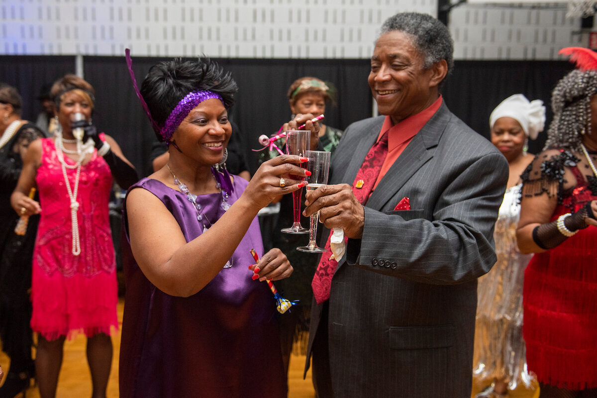In a decorated gymnasium, two seniors in 1920s party outfits toast their glasses of punch while smiling. There are more revelers behind them in flapper dresses, long beaded necklaces, and 1920s headbands.