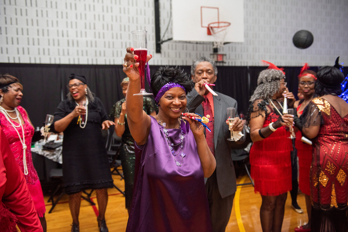 In a decorated gymnasium, seniors in 1920s-era party costumes dance and raise their plastic champagne flutes in the air. Several are blowing paper horns and noisemakers typical of New Years Eve parties.