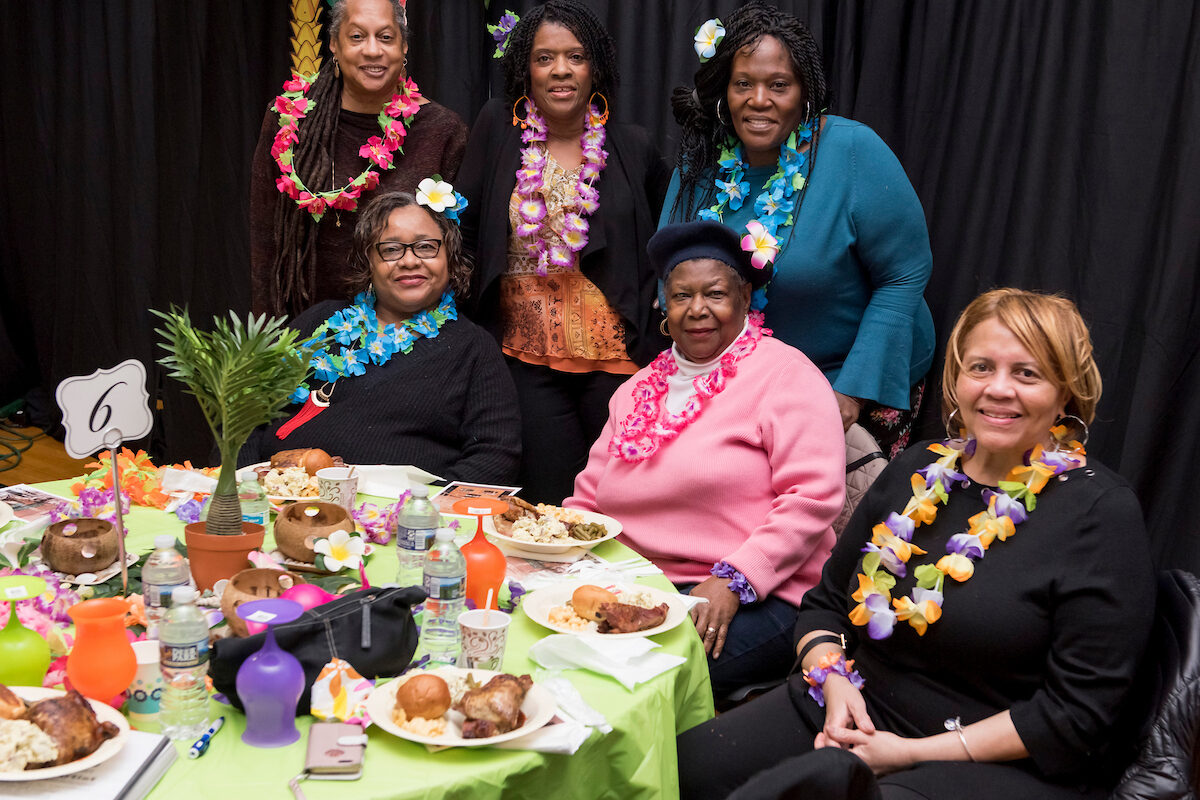 Six women wearing colorful flower lei necklaces smile for a photo around a table covered with plates of food and Hawaiian luau-style decorations. Many of the women also have a large flower behind one ear.