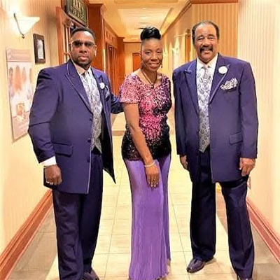 Two men in matching purple suits and ties stand on either side of a woman in a purple floor-length gown.