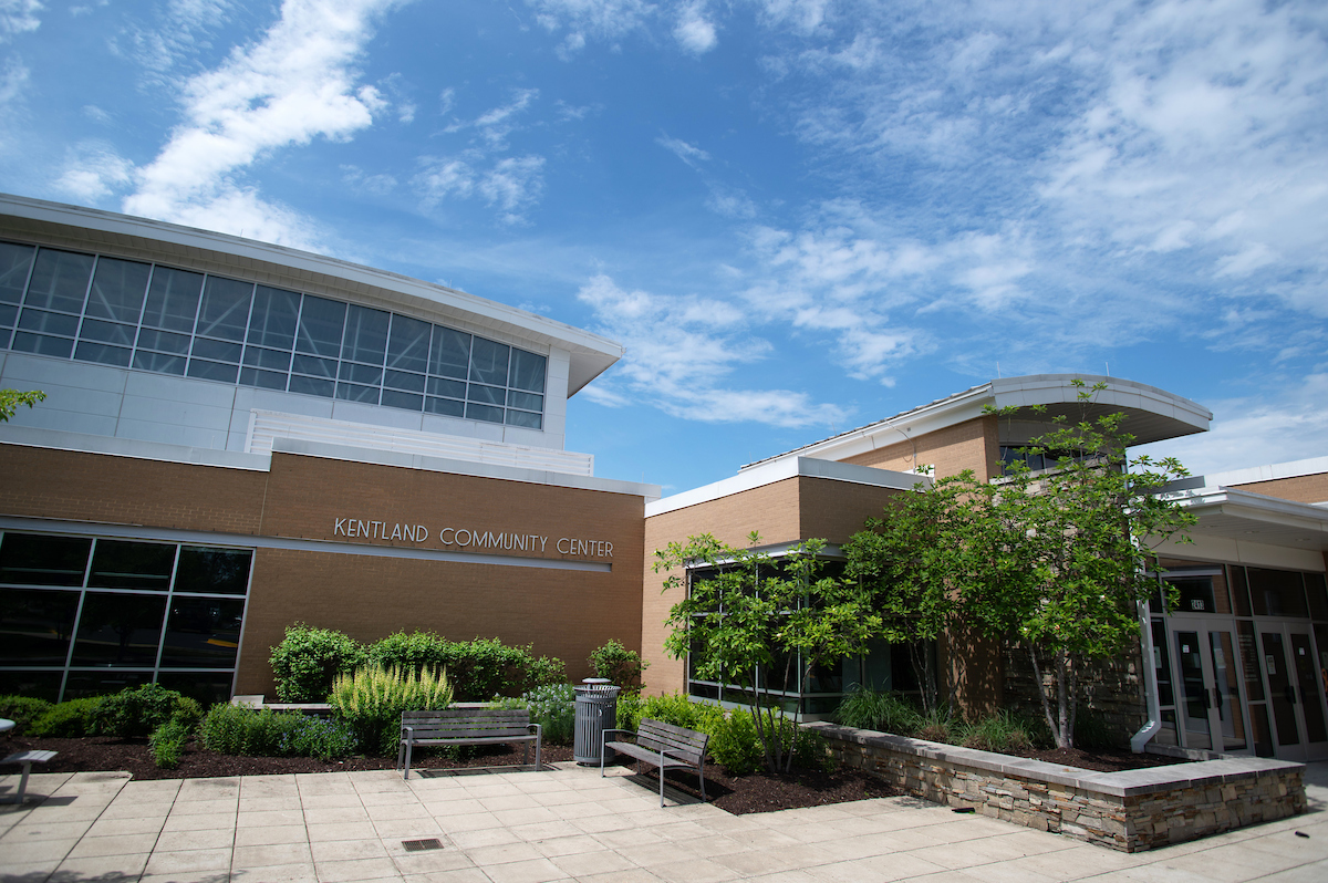 A large brick, glass, and metal building with landscaped planters full of trees and foliage. Several benches stand near the plants. Letters on the building read KENTLAND COMMUNITY CENTER.