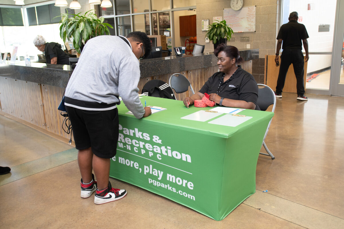 A smiling woman sits behind a check-in table covered with a green Parks and Rec tablecloth in a lobby, next to the reception desk. A man stands at her table filling out a form.