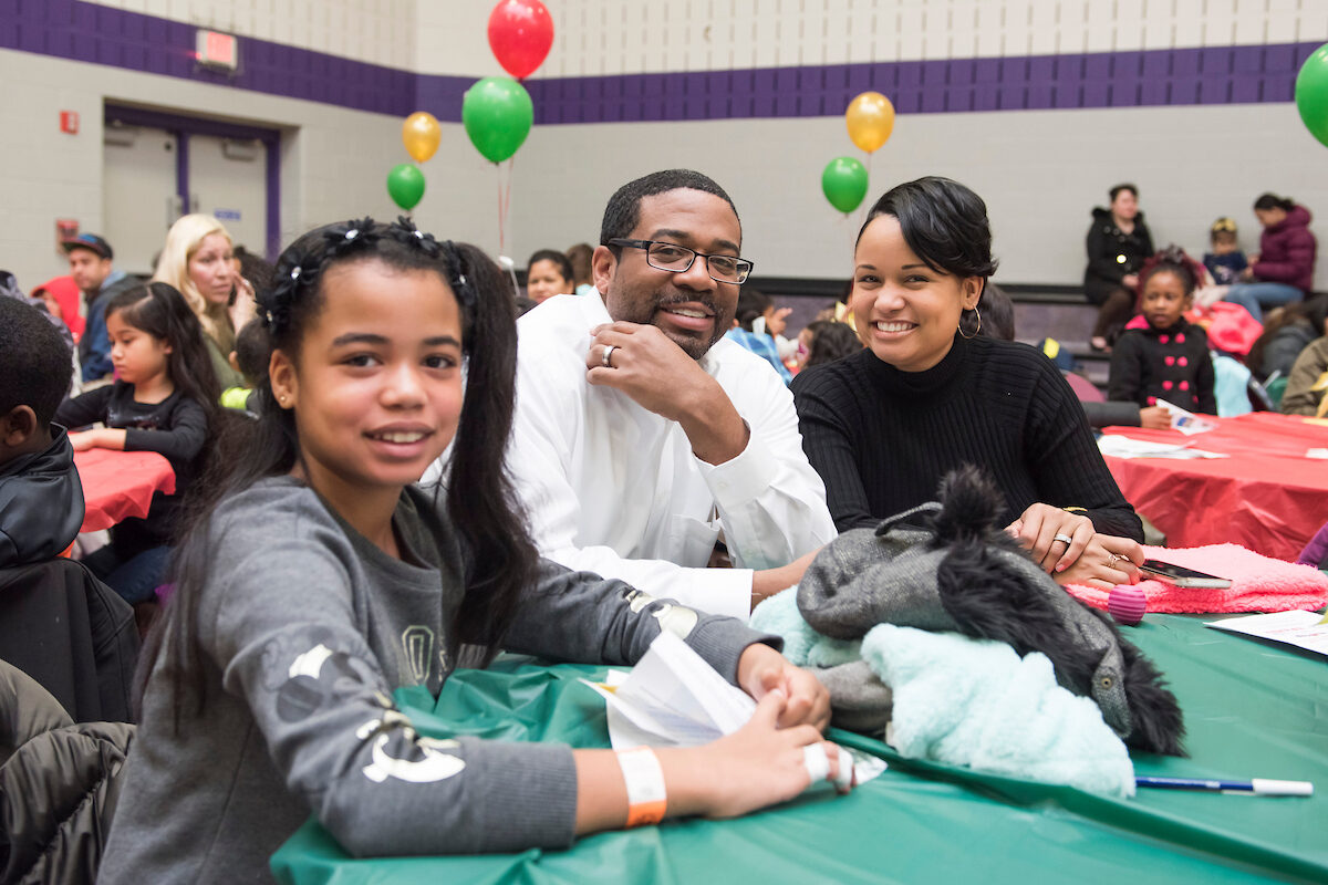 Inside a gymnasium with purple trim, decorated with colorful balloons, a family seated at a table smiles for the camera. Behind them are other tables, all with colorful tablecloths, filled with children and families.