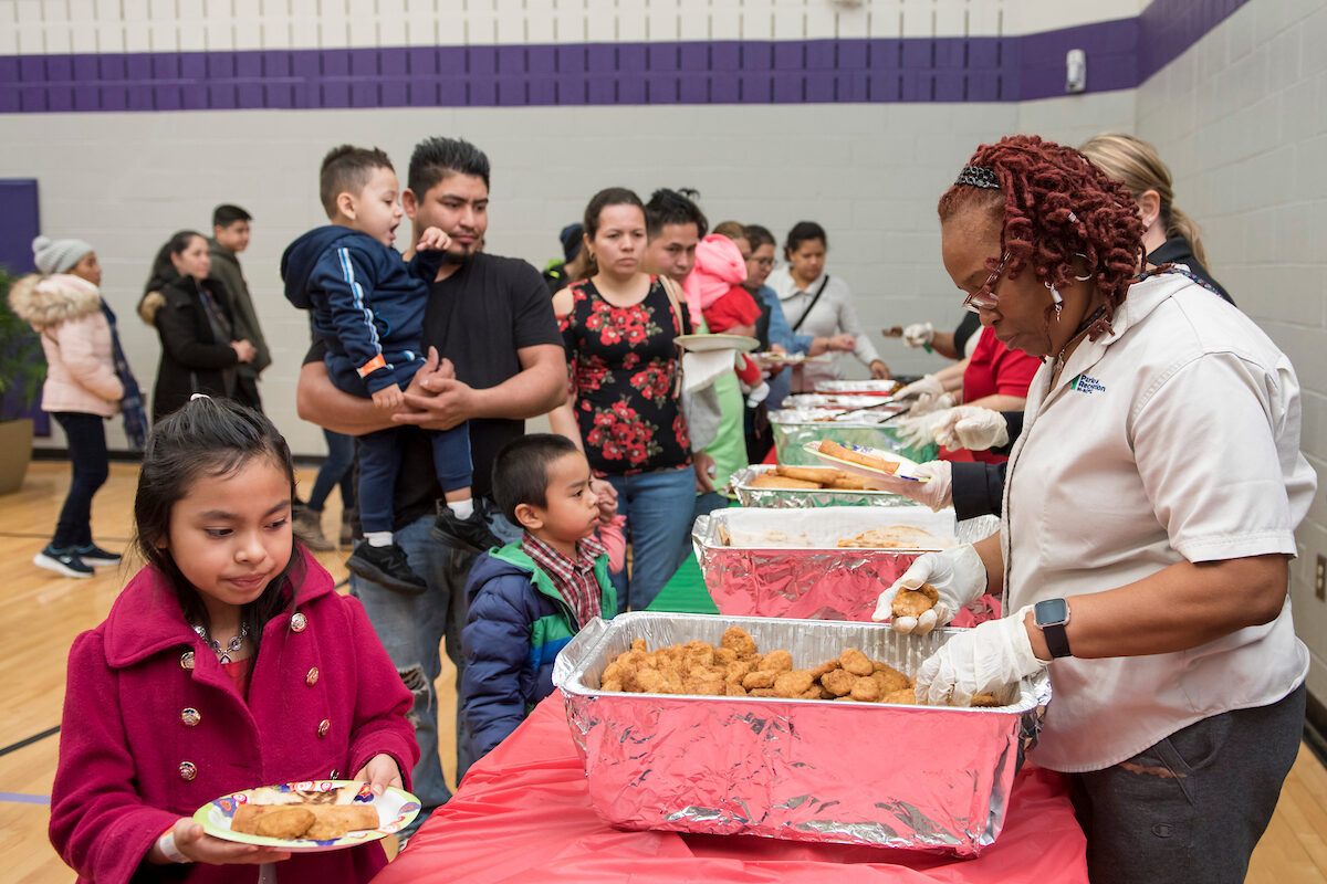 Inside a gymnasium with purple trim, adults in tan staff shirts stand behind long tables serving food from banquet dishes to a line of customers. The customers are mainly parents and small children, some wearing winter coats.